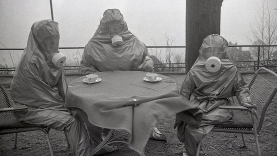 Drei Personen sind in ABC-Schutzanzügen und Atemschutzmasken an einem Tisch. © Staatsarchiv Hamburg Plankammer 