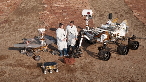 Zwei Raumfahrtingenieure stehen zwischen drei Generationen Marsrovern auf einem Testgelände in Pasadena - darunter Curiosity. © NASA/JPL-Caltech 