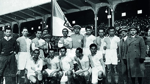 Eine Fußballmannschaft posiert auf einem historischen Foto mit Fahnen und Stadiontribünen im Hintergrund. © Anne Siegel 