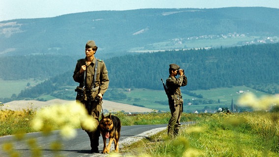 Zwei Grenzer der DDR-Grenztruppen kontrollieren 1987 mit einem Schäferhund einen Abschnitt der innerdeutschen Grenze am Brocken. © picture-alliance/ ZB Foto: Hans Wiedl