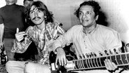 Der britische Musiker und Beatle George Harrison (l.) lauscht am 3.8.1967 bei einem Treffen in Hollywood dem indischen Musiker Ravi Shankar, der auf einer Sitar (indische Langhalslaute) spielt. © picture-alliance / dpa | UPI 