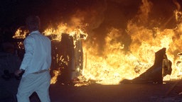 Ein Mann steht am 27. August 1992 vor brennenden Pkw auf einer Straße am Asylbewerberheim in Rostock-Lichtenhagen. © picture alliance / ZB Foto: Bernd Wüstneck