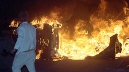 Ein Mann steht am 27. August 1992 vor brennenden Pkw auf einer Straße am Asylbewerberheim in Rostock-Lichtenhagen. Ein Mann steht am 27. August 1992 vor brennenden Pkw auf einer Straße am Asylbewerberheim in Rostock-Lichtenhagen. © picture alliance / ZB Foto: Bernd Wüstneck