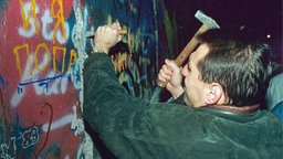 Ein Mann haut mit Hammer und Meißel Stücke aus der Berliner Mauer heraus. © picture alliance / dpa 