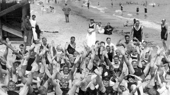 Viele fröhliche Menschen am Strand von Usedom heben ihre Füße in die Luft. Historische Aufnahme um 1920. © picture alliance / imageBROKER Foto: Rosseforp