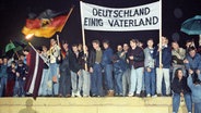 Mit der Deutschlandfahne und einem Transparent "Deutschland Einig Vaterland" stehen zahlreiche Berliner am 22. Dezember 1989 auf der Berliner Mauer am Brandenburger Tor. © picture-alliance/ dpa | DB dpa 