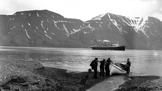 Historische Aufnahme des Dampfschiffs "Augusta Victoria" der Reederei Hapag in einem Fjord mit Personen im Vordergrund.  