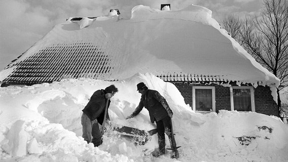 Zwei Männer stehen im Schneewinter 1978/79 vor einem Haus im Kreis Rendsburg-Eckernförde und versuchen ein Auto zu befreien, das komplett von einem Schneeberg bedeckt ist. © picture alliance/dpa/Werner Schilling Foto: Werner Schilling