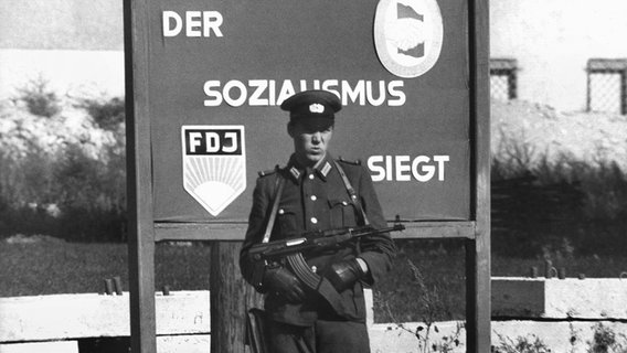 Ein bewaffneter Volkspolizist steht vor einem Schild mit der Propagandaparole "Der Sozialismus siegt". © picture-alliance / dpa 
