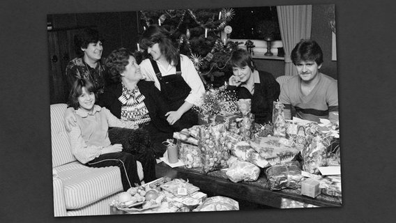 Eine Familie sitzt am Heiligabend im Wohnzimmer zusammen und freut sich über viele Weihnachtsgeschenke auf dem Tisch. © imago/Werner Otto Foto: Werner Otto
