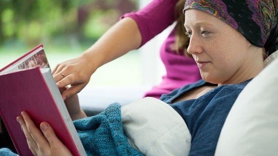 Ein Mädchen mit einem Kopftuch liegt in einem Krankenbett und bekommt etwas in einem Buch gezeigt. © PantherMedia Foto: photographee.eu
