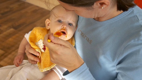 Baby mit Lätzchen wird von seiner Mutter mit Karottenbrei gefüttert © imago/INSADCO 