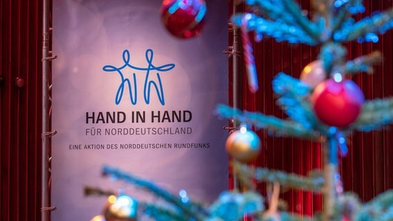 Das Logo der Aktion "Hand in Hand", im Vordergrund ein geschmückter Weihnachtsbaum. © NDR Foto: Axel Herzig