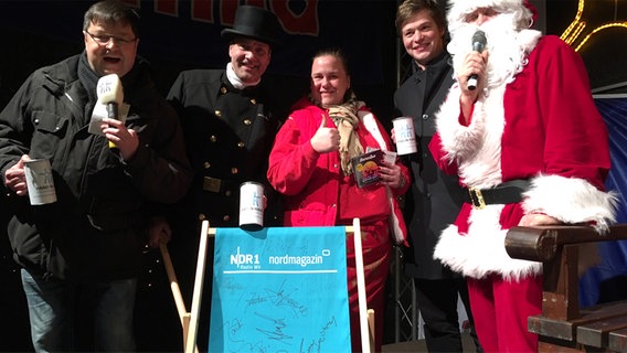 Leif Tennemann steht gemeinsam mit einem Schornsteinfeger, einer Frau, einem Mann und einem Weihnachtsmann vor einem NDR Liegestuhl.  