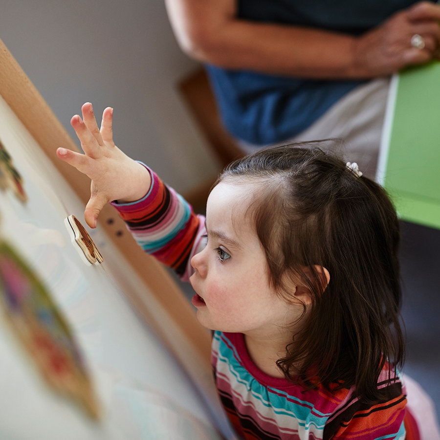 Ein kleines Mädchen klebt einen Sticker an eine Tafel, im Hintergrund sitzt eine Frau. © Lebenshilfe Foto: David Maurer