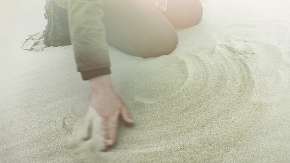 Ein knieender Mensch wischt Spuren in weißem Sand. © phootocase.de Foto: lichtsicht