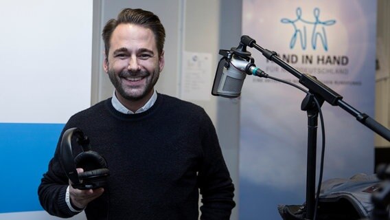 Dennis Wilms steht mit Kopfhörern in der Hand neben einem Mikrofon im Hörfunkstudio. © NDR Foto: Dominik Dührsen