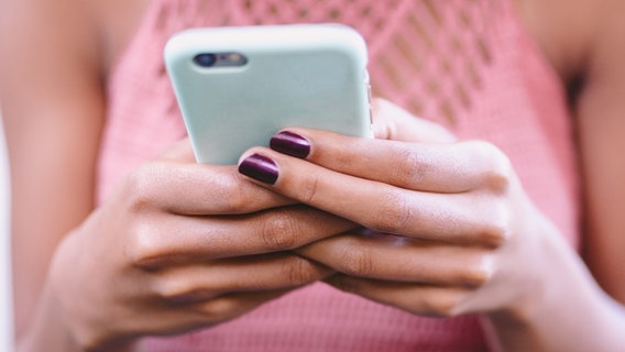 Ein Handy wird von einer jungen Frau, die violetten Nagellack und sommerliche Kleidung trägt in den Handen gehalten. © criene Foto: criene