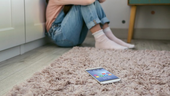 Symbolfoto: Eine junge Frau hockt in einer Zimmerecke, auf dem Boden liegt ein Smartphone. © davidpereiras / Photocase Foto: davidpereiras