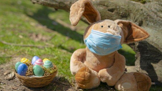 Eine Osterhasenpuppe mit Mundschutz sitzt neben einem Körbchen mit Ostereiern. © picture alliance Foto: Jens Krick, flashpic