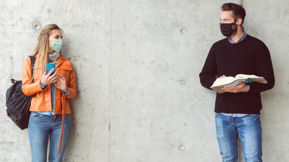 Zwei junge Menschen mit Maske stehen an einer Betonwand mit Büchern und Smartphone in der Hand © Colourbox Foto: Kzenon