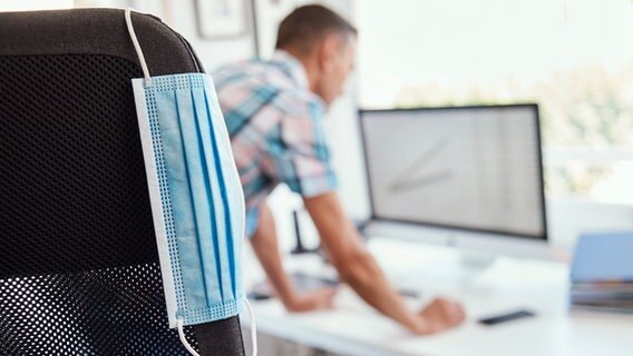 Eine Einmalschutzmaske hängt an einem Bürostuhl, im Hintergrund ist ein Mann am Computer zu sehen. © Colourbox Foto: nito