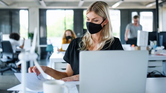 Eine Frau mit Schutzmaske arbeitet in einem Büro mit mehreren Angestellten. © Colourbox 