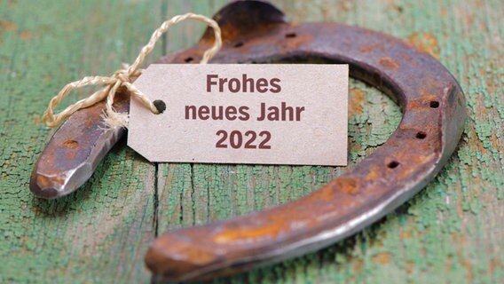 Eine rostiges Hufeisen liegt mit einem Schild auf dem "Frohes neues Jahr 2022" steht auf einem hölzernen Untergrund. © Colourbox 