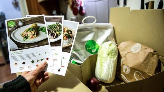 Eine Person hält drei Rezeptkarten vor den Inhalt einer Kochbox © picture alliance / dpa Foto: Daniel Naupold