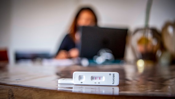 Auf einem Tisch liegt ein positiver Corona-Schnelltest. Im Hintergrund sieht man unscharf eine Frau am Laptop. © picture alliance / Zoonar Foto: Robert Kneschke