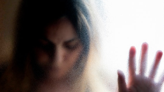 Eine Frau stützt sich mit gesenktem Blick mit der Hand gegen eine krauselige Glasscheibe. © Imago Images / Westend61 Foto: Westend61