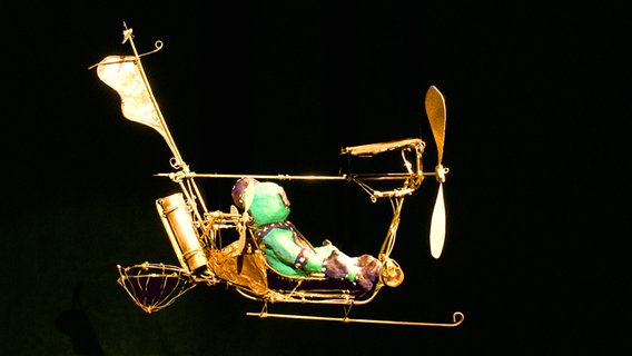Ein goldenes Fahrzeug mit einer grünen Figur ist im Theater vor einer schwarzen Wand zu sehen. © NDR Foto: Linda Ebener