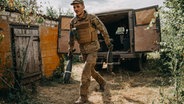 Ein Soldat in ukrainischer Uniform trägt in jeder Hand eine Mörsergranate. Er entfernt sich von einem Militärtransporter mit geöffneten Hecktüren. © picture alliance / AA Foto: Wojciech Grzedzinski