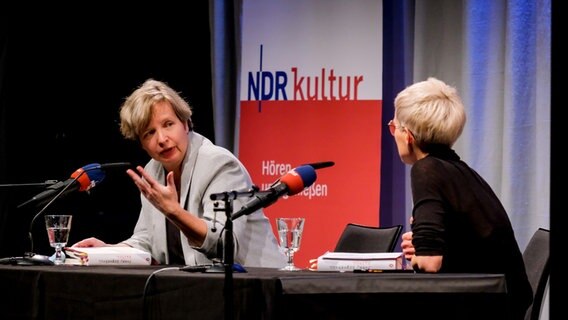 Jenny Erpenbeck und Alexandra Friedrich sitzen an einem Tisch auf einer Bühne und schauen sich an. © Dietrich Kühn Foto: Dietrich Kühne