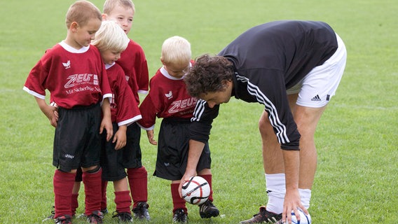 Ein Fußballtrainer bringt vier kleinen Jungen das Schießen bei. Themenbild für Engagement im Verein. © imago/JOKER Foto: RalfxGerard