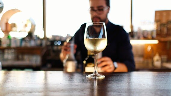 Ein Glas Wein steht auf einem Tresen. Im Hintergrund steht ein Barkeeper. © picture alliance / Westend61 Foto: Angel Santana Garcia
