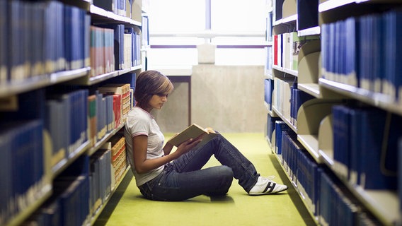 Junge Frau sitzt zwischen den Bücherregalen in der Bibliothek und liest in einem Buch. © picture alliance / imageBROKER | photorevolution 
