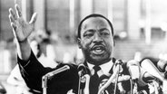 Der Bürgerrechtler Martin Luther King hebt eine Hand hoch. © picture alliance/Glasshouse Images 