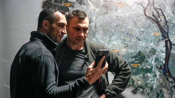Vitali Klitschko (r), Bürgermeister von Kiew und ehemaliger Box-Profi, und sein Bruder Wladimir Klitschko, ebenfalls ehemaliger Box-Profi, schauen auf ein Smartphone im Rathaus in Kiew. © Efrem Lukatsky/dpa/AP Foto: Efrem Lukatsky