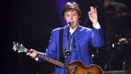 Paul Mc Cartney steht mit einer Gitarre in der Hand auf einer Bühne und hebt die Hand. © dpa Foto: O Corsan