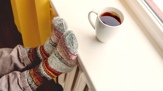 Eine Person sitzt mit Socken vor einer Heizung. Auf der Fensterbank steht eine Tasse Tee. © picture alliance / PantherMedia Foto: Ronalds Stikans