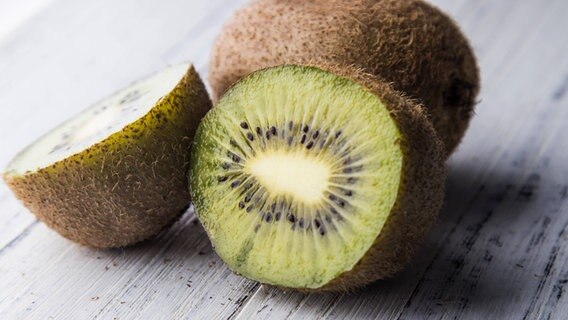Eine aufgeschnittene und eine ganz Kiwi liegen auf einem hellen Holzuntergrund. © imago Foto: ZUMA Press