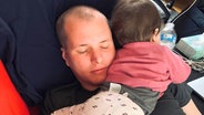 Washington Korrespondent Torben Ostermann liegt mit geschlossenen Augen auf dem Sofa und hält sein Baby im Arm. © ARD Foto: Torben Ostermann