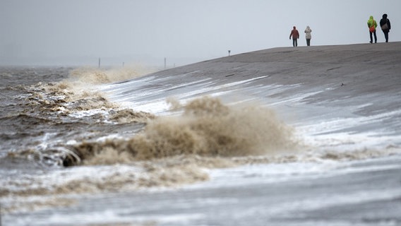 Spaziergänger laufen auf dem Deich in Norddeich, an den die Wellen schlagen. © picture alliance Foto: Matthias Balk