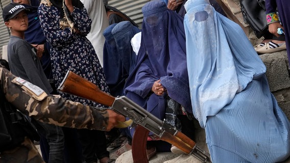 Ein Taliban-Kämpfer in Kabul hält eine Kalaschnikow, während im Hintergrund Frauen in Burkas sitzen. © picture alliance/dpa/AP Foto: Ebrahim Noroozi