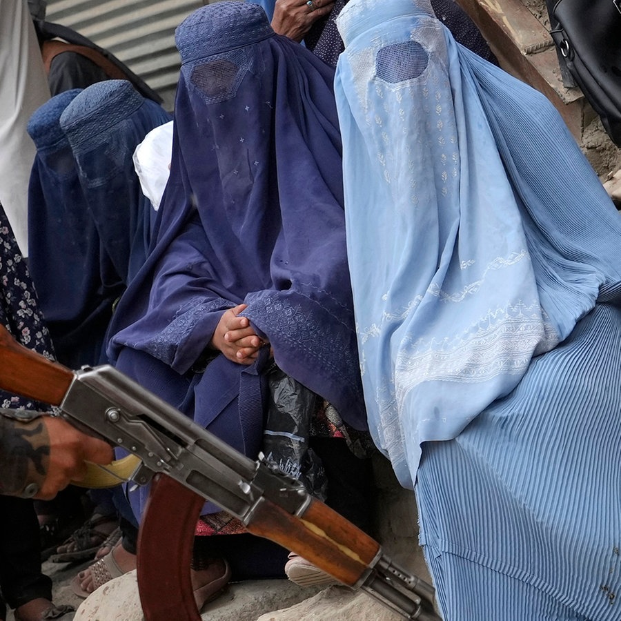 Ein Taliban-Kämpfer in Kabul hält eine Kalaschnikow, während im Hintergrund Frauen in Burkas sitzen. © picture alliance/dpa/AP Foto: Ebrahim Noroozi