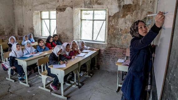 Ein Klassenzimmer in Afghanistan: Eine Lehrerin schreibt etwas an die Tafel, während im Hintergrund Mädchen an Schreibtischen sitzen. © picture alliance / ASSOCIATED PRESS Foto: Ebrahim Noroozi