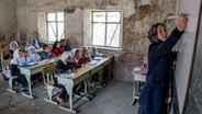Ein Klassenzimmer in Afghanistan: Eine Lehrerin schreibt etwas an die Tafel, während im Hintergrund Mädchen an Schreibtischen sitzen. © picture alliance / ASSOCIATED PRESS Foto: Ebrahim Noroozi