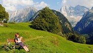 Frau liest ein Buch auf einer Bank - im Hintergrund eine Bergkulisse © picture alliance / imageBROKER | Oliver Gerhard 