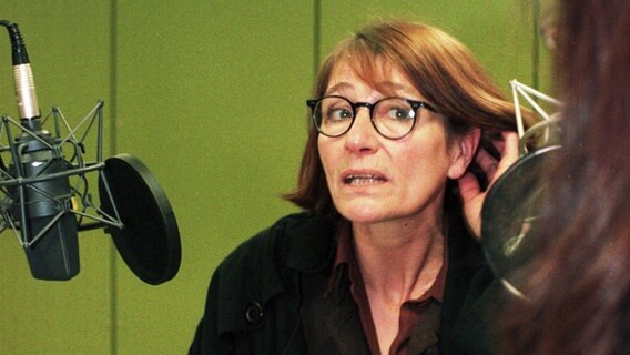 Eine rothaarige Frau mittleren Alters steht in einem Studio vor einem Mikrofon und spricht hinein. Sie hält ihre Hand am Ohr als würde sie lauschen. © NDR 